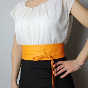 May include: A vibrant orange leather obi belt tied with a bow. The belt is worn over a black skirt and a white blouse. The belt's bright colour provides a striking contrast against the darker clothing.