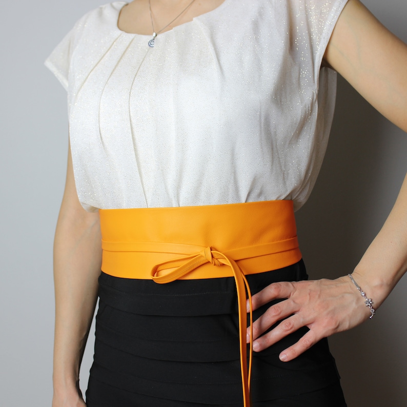 May include: A vibrant orange leather obi belt tied with a bow. The belt is worn over a black skirt and a white blouse. The belt's bright colour provides a striking contrast against the darker clothing.