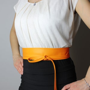 May include: A bright orange leather belt tied in a bow, worn over a black skirt and a white top. The belt's colour contrasts with the black skirt. The bow adds a decorative touch. The outfit is simple and elegant.