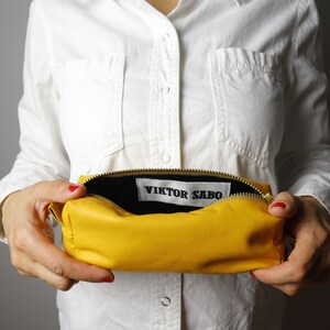 May include: A vibrant yellow leather pouch with a gold zip, held open to show a black interior. The label inside reads "VIKTOR SABO". The rectangular pouch is likely made of soft leather.