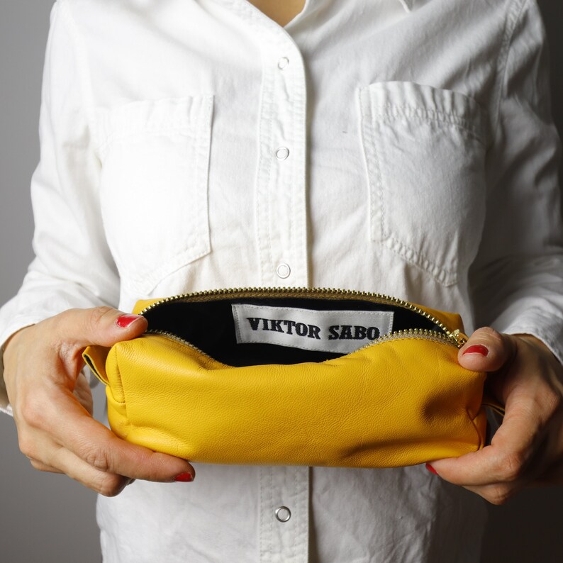 May include: A vibrant yellow leather pouch with a gold zip, held open to show a black interior. The label inside reads "VIKTOR SABO". The rectangular pouch is likely made of soft leather.