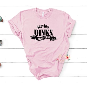 Könnte beinhalten: Rosa T-Shirt mit schwarzem Text, der "Serving Dinks Since 1958" mit einem schwarzen Bandbanner lautet.