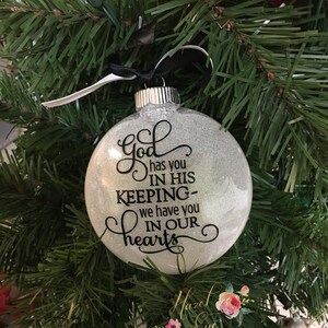 May include: A silver glitter Christmas ornament with the text "God has you in his keeping - we have you in our hearts".