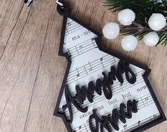 Op de afbeelding: Zwarte houten kerstboomversiering met de woorden "Amazing Grace" in cursief. De versiering heeft een achtergrond van muzieknoten en is versierd met een zwart-wit geruit lint.