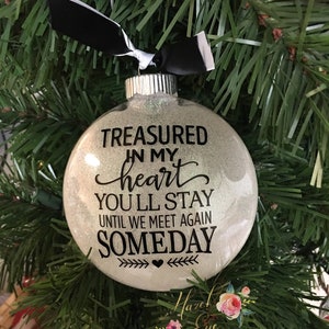 May include: A white glitter Christmas ornament with black lettering that reads "Treasured in my heart you'll stay until we meet again someday" with a heart and arrows on the bottom.