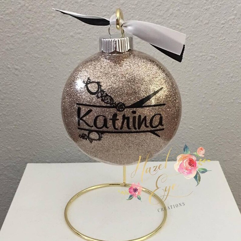 Op de afbeelding: Een gouden glitter kerstbal met een zwart silhouet van een schaar en de naam "Katrina" erop. De bal staat op een gouden standaard.