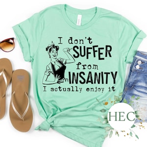 Puede incluir: Una camiseta verde claro con una ilustración de estilo retro de una mujer con una expresión de sorpresa. El texto en la camiseta dice "No sufro de locura, en realidad la disfruto".