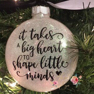 May include: White glitter Christmas ornament with black lettering that reads "It takes a big heart to shape little minds".