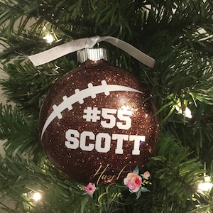 May include: A brown glitter football-shaped Christmas ornament with white stitching detail. The ornament features the text "#55 SCOTT" in white. A white ribbon is attached for hanging. The ornament is displayed on a Christmas tree.