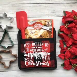 May include: A Christmas-themed gift set featuring a red spatula, cookie cutters, and a black and red plaid oven mitt with the text "Just Rollin' By To Wish You a Merry Christmas". A package of Betty Crocker cookie mix is also included.
