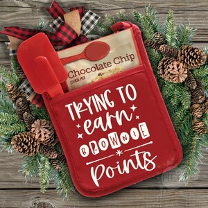 Puede incluir: Manopla de horno roja con el texto "Trying to earn brownie points" en blanco. Un paquete de mezcla para galletas con chispas de chocolate está en el bolsillo de la manopla.