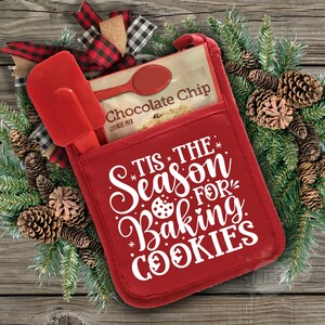 Könnte beinhalten: Ein roter Ofenhandschuh mit dem Text "Tis the season for baking cookies" in Weiß. In der Tasche des Handschuhs befindet sich eine Packung Schokoladen-Chip-Kekse.