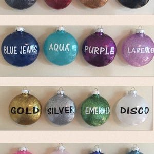Op de afbeelding: Een verzameling van 16 ronde kerstballen met glitterafwerking. De ballen zijn versierd met verschillende kleuren en woorden, waaronder gunmetal, crimson, copper, black, blue jeans, aqua, purple, lavender, gold, silver, emerald, disco, pink ice, soft pink, royal blue en light blue.
