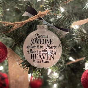 May include: White glitter Christmas ornament with the text "Because someone we love is in Heaven, There's a little bit of Heaven in our home."  The ornament is hanging on a Christmas tree.