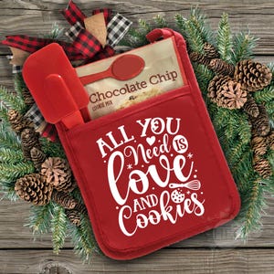 Może przedstawiać: Czerwona rękawica do piekarnika z napisem "All you need is love and cookies" w kolorze białym. W kieszeni rękawicy znajduje się opakowanie mieszanki do ciasteczek z kawałkami czekolady i czerwona szpatułka.
