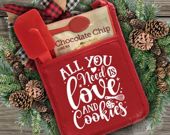 Op de afbeelding: Een rode ovenwant met de tekst "All you need is love and cookies" in wit. Een pakje chocolade chip koekjesmix en een rode spatel zitten in de zak van de ovenwant.