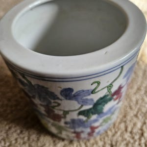 May include: A white ceramic planter with a wide rim and a painted floral design. The planter features a blue, green, and red grape vine pattern around the exterior. The interior is white and the planter sits on a beige surface.