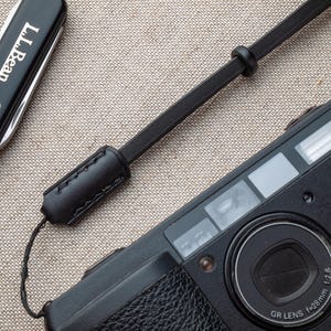 Może przedstawiać: Czarny aparat fotograficzny z paskiem skórzanym i czarnym scyzorykiem z logo L.L.Bean. Obiektyw aparatu oznaczony GR LENS f=28mm 1:2.8. Pasek jest przymocowany do aparatu i ma przeszyty skórzany detal.