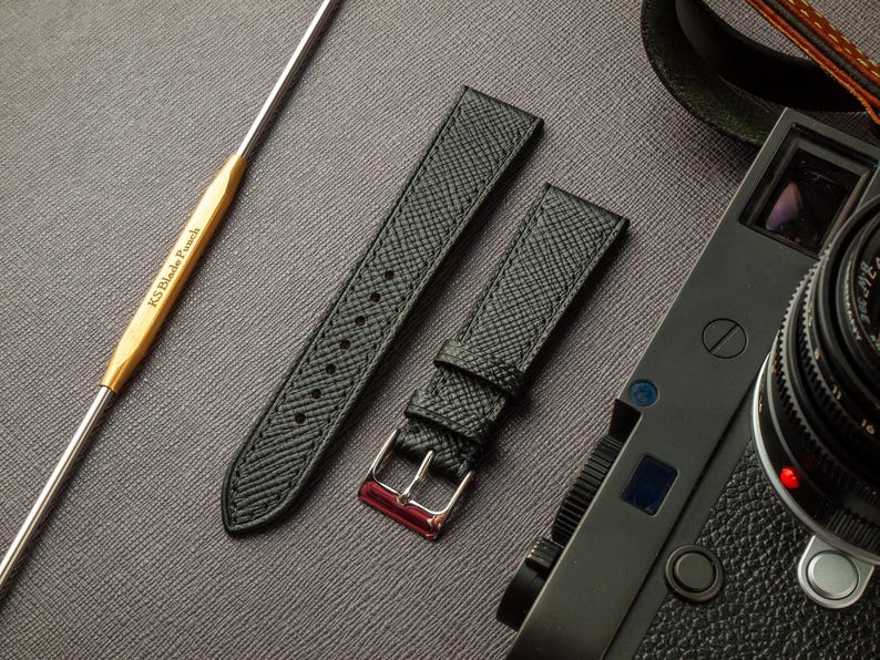 May include: Two black leather watch straps with a textured finish. The straps have a silver buckle and a red accent. The straps are laid out on a gray surface.