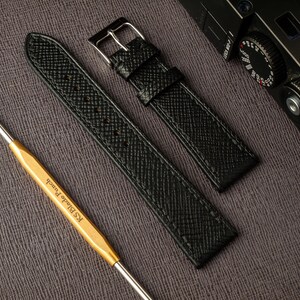 May include: Two black leather watch straps with a textured finish. The straps are laid out on a gray surface. One strap has a silver buckle.
