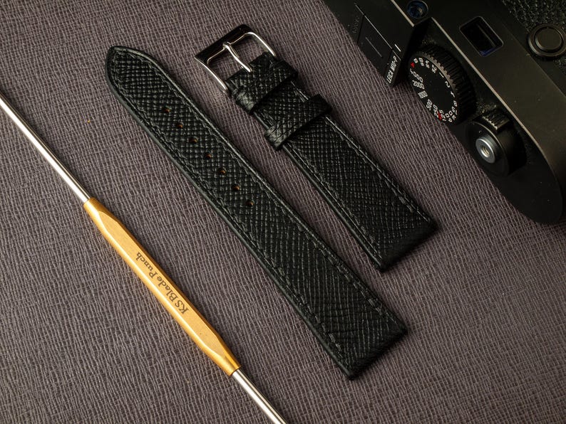 May include: Two black leather watch straps with a textured finish. The straps are laid out on a gray surface. One strap has a silver buckle.