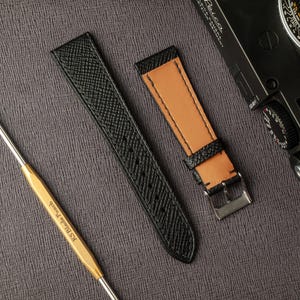 May include: Two watch straps, one black and one brown, with a stitching detail. The straps are laid out on a gray textured surface.