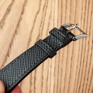 May include: A black leather watch strap with a silver buckle. The strap has a textured surface.