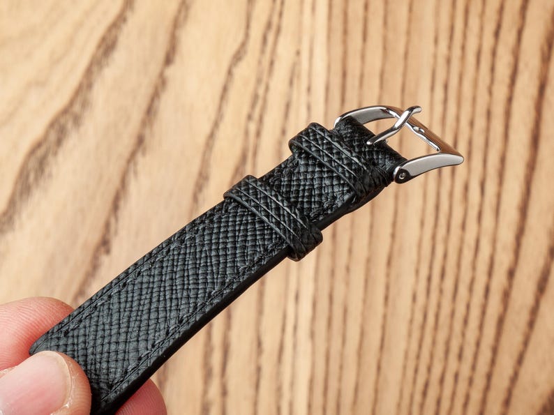 May include: A black leather watch strap with a silver buckle. The strap has a textured surface.