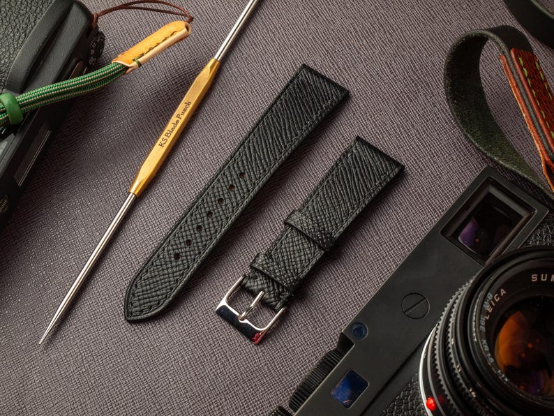 May include: Two black leather watch straps with a textured finish. The straps have silver buckles and are laid out on a gray surface. The straps are next to a camera and a leather strap.