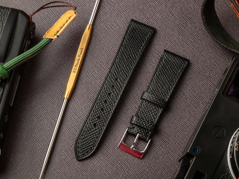 May include: Two black leather watch straps with a textured finish. The strap on the left has a pointed end, while the strap on the right has a buckle with a red accent.