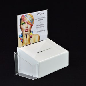 May include: White ballot box with a slot for suggestions or collections. A clear acrylic pocket is attached to the side. A sign holder displays a colourful image and text: "Suggestion / Collection Ballot Box" and "A5 Landscape Poster Holder".