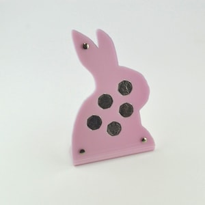 May include: A pink bunny-shaped coin holder with five coin-sized holes and gold-coloured rivets. The bunny has a simple, stylised design, and the coins are visible within the holes. The holder is made of a smooth, solid material.