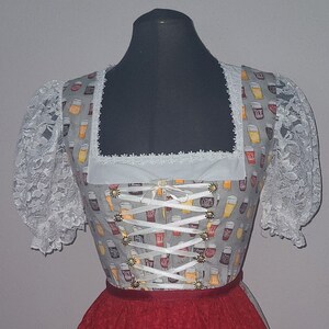 Op de afbeelding: Een witte dirndl met een grijs lijfje met een herhalend patroon van bierglazen. Het lijfje heeft een witte kantversiering en een wit lint dat in het midden is gebonden tot een strik. De rok is rood met een witte en gele rand.