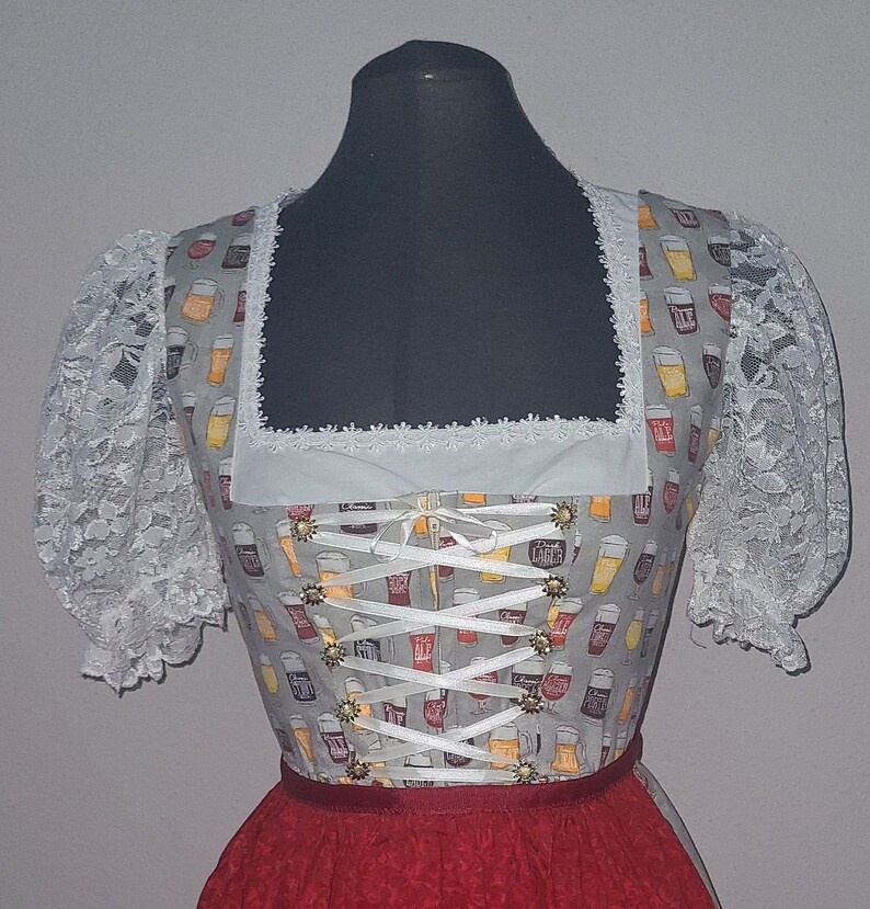 Op de afbeelding: Een witte dirndl met een grijs lijfje met een herhalend patroon van bierglazen. Het lijfje heeft een witte kantversiering en een wit lint dat in het midden is gebonden tot een strik. De rok is rood met een witte en gele rand.