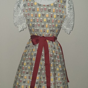 Op de afbeelding: Een grijze dirndl-jurk met een wit kantboordje en een rode strik. De jurk heeft een patroon van bierglazen met verschillende soorten bier.