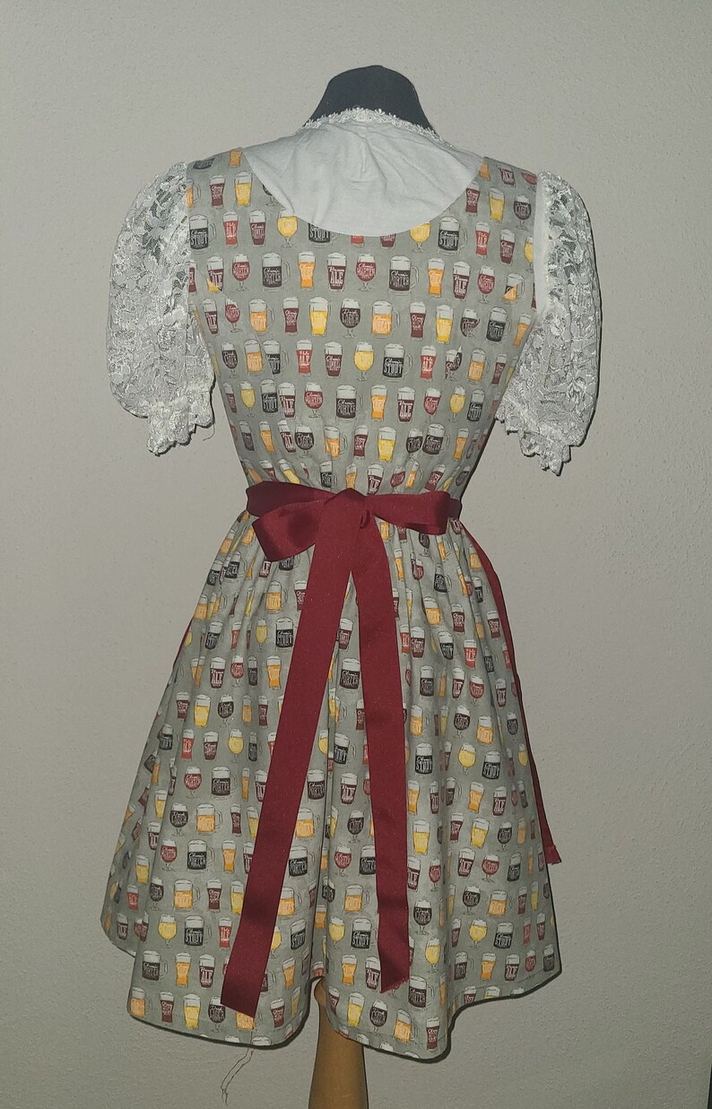 Op de afbeelding: Een grijze dirndl-jurk met een wit kantboordje en een rode strik. De jurk heeft een patroon van bierglazen met verschillende soorten bier.