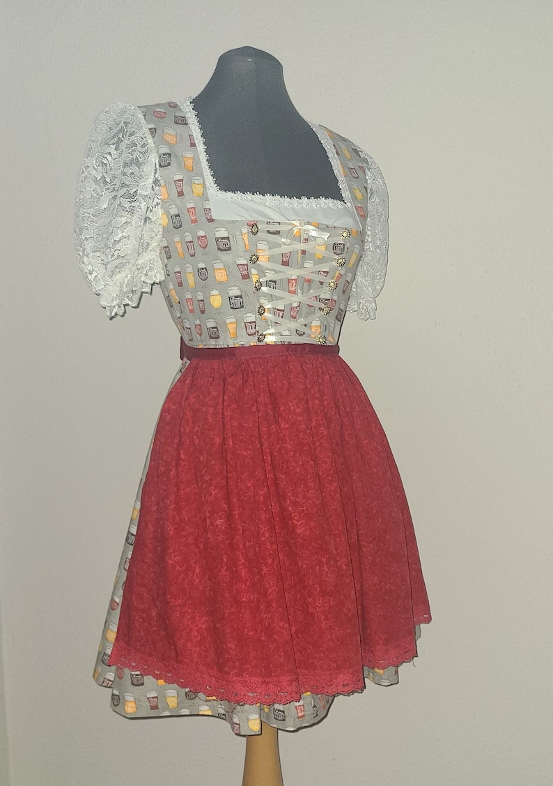 Op de afbeelding: Een wit en grijs dirndl met een rode rok. Het lijfje is versierd met een patroon van bierglazen. De rok heeft een kantversiering.