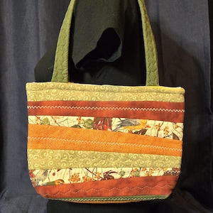 May include: A quilted tote bag with a patchwork design. The bag features horizontal stripes in shades of orange, red, and green, with a floral and bird pattern. The handles are a quilted olive green. The bag is approximately 30 cm wide.