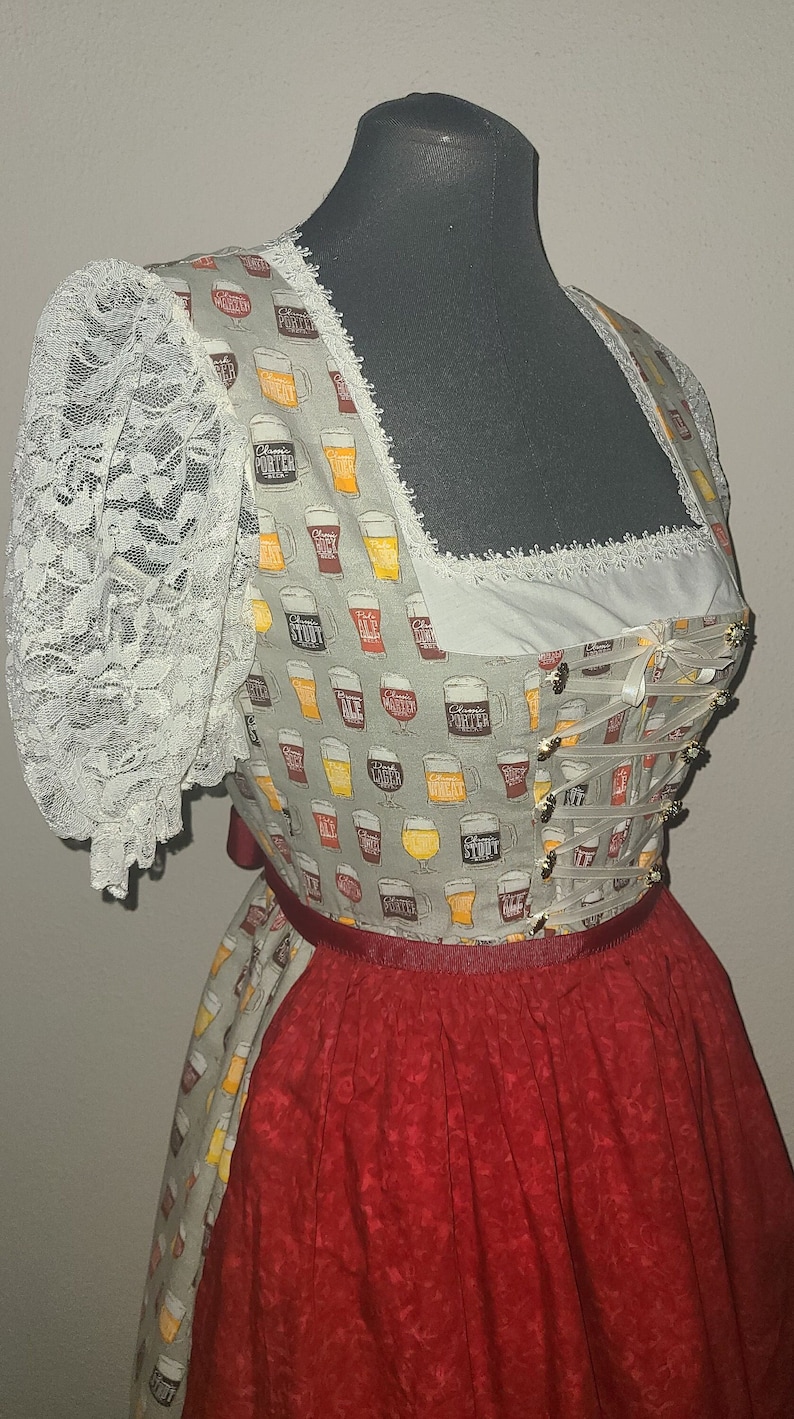 Op de afbeelding: Een traditionele dirndl jurk met een wit kant corsage en een grijze rok met een bierglas print. De rok heeft een rode tailleband en een rode onderrok.