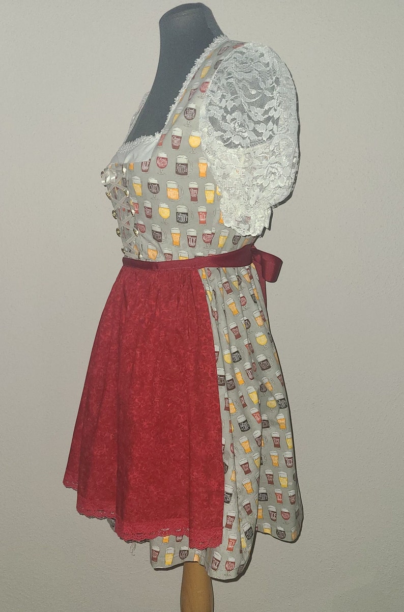 Op de afbeelding: Een grijs en wit dirndl met een rode rok en een kantboord. Het boord is versierd met een patroon van bierglazen. De rok heeft een kantbies.