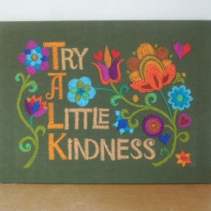 May include: Embroidered artwork on a green canvas. The text "TRY A LITTLE KINDNESS" is stitched in orange thread, surrounded by colourful floral designs in blue, orange, purple, and pink. A decorative piece for home decor.