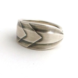 May include: A silver ring with a geometric design. The ring features a series of parallel lines and angular shapes, creating a textured appearance. The ring is set against a white background.