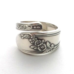May include: A silver spoon ring with floral detailing. The ring is made from a spoon, with the handle wrapped around to form the band. The spoon's bowl features an embossed floral design. The ring is set against a white background.