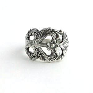 May include: A silver-toned ring with an ornate floral design. The ring features a central flower and swirling leaf patterns. The metal has a slightly aged appearance, and the ring is set against a white background.