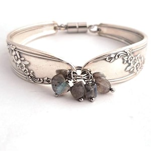 May include: A silver bracelet crafted from repurposed silverware, featuring floral detailing and a magnetic clasp. The bracelet is adorned with a cluster of small, gray gemstone beads, adding a touch of elegance to the unique design.