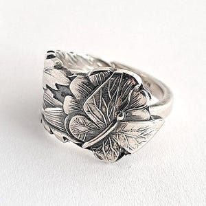 May include: A silver ring with a detailed floral design. The ring features a prominent leaf and flower motif, with intricate veining and textures. The ring is set against a white background, highlighting the metallic sheen and craftsmanship.