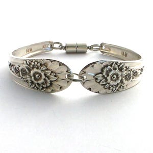 May include: A silver bracelet crafted from repurposed silverware. The bracelet features two spoon heads with floral designs, connected by a magnetic clasp. The spoon heads are adorned with detailed floral patterns, creating a vintage aesthetic.