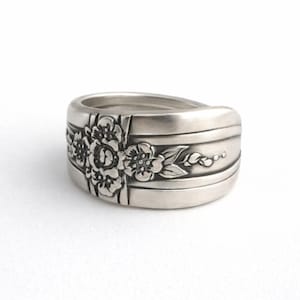 May include: A silver spoon ring with a floral design. The ring features a detailed flower and leaf pattern on the front, with horizontal lines on the band. The ring is made of a shiny, metallic material.