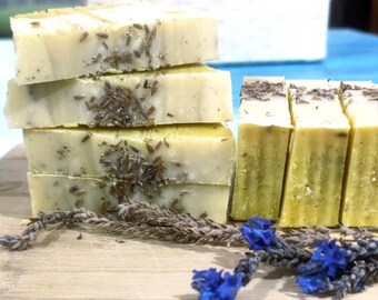 Puede incluir: Una pila de cuatro barras de jabón hechas a mano con lavanda y una ramita de flores de lavanda sobre una tabla de cortar de madera.