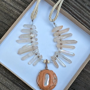 Peut inclure: Un collier composé de cristaux de quartz transparents et d'un pendentif enroulé de fil de cuivre avec une base en argile brune et une coquille irisée blanche. Le collier est sur un fond beige.